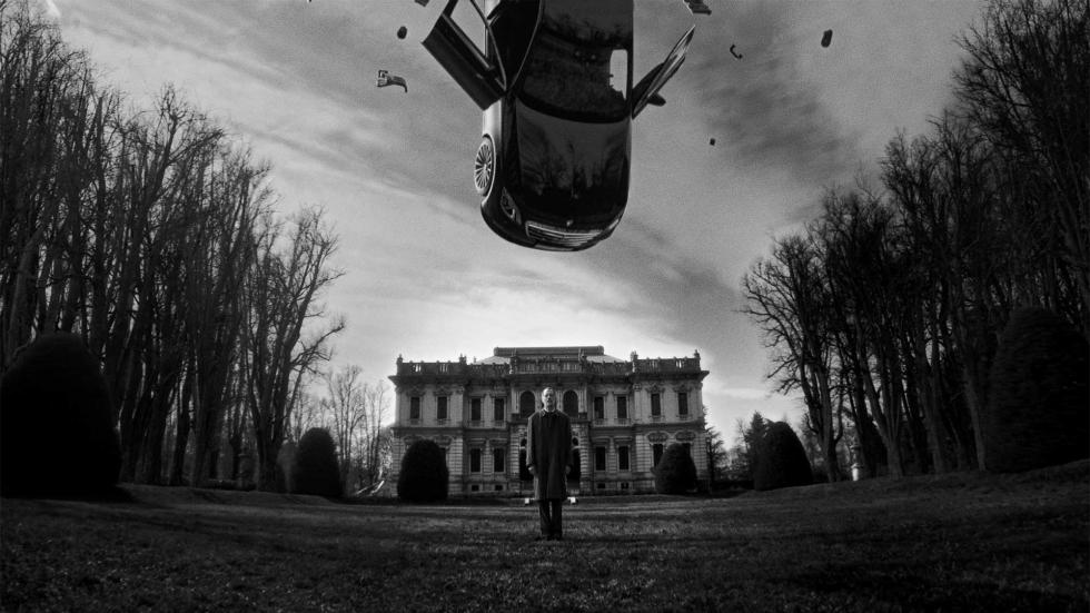 A car hanging in the sky above a man standing with a mansion behind him