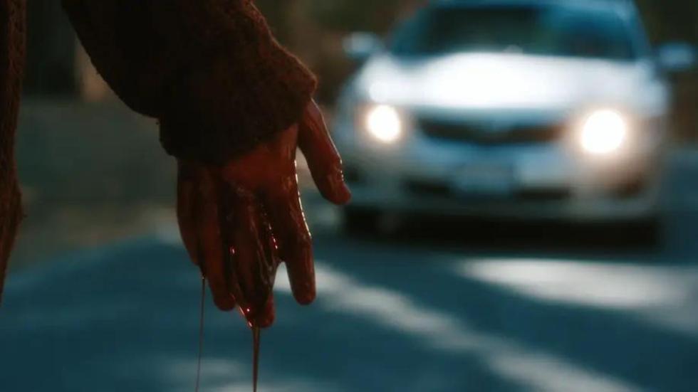 A bloody hand drips blood on the road as a car approaches