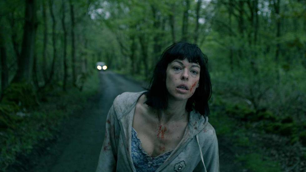 White Settlers bloodied woman limps down dirt road with car lights in the background